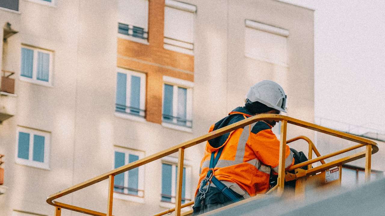 Technicien en casque blanc et veste haute visibilité orange sur une nacelle, le long d’une façade d’immeuble claire