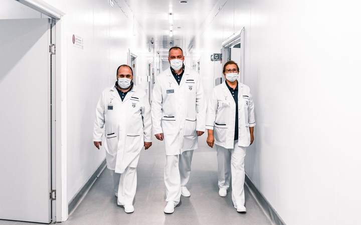Trois soignants masqués en blouse blanche marchant dans un couloir d’hôpital