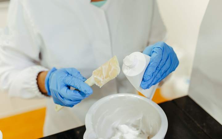 Préparation d’une crème en laboratoire : mains gantées tenant une spatule et un flacon souple au-dessus d’un bol de mélange