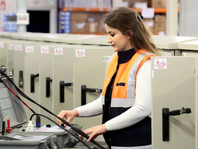 Technicienne en gilet orange réalisant des mesures avec un appareil de test devant des armoires électriques