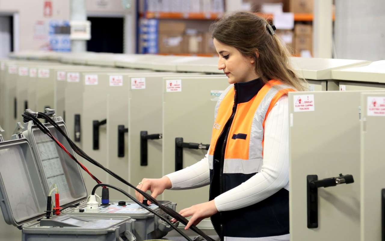 Technicienne en gilet orange réalisant des mesures avec un appareil de test devant des armoires électriques