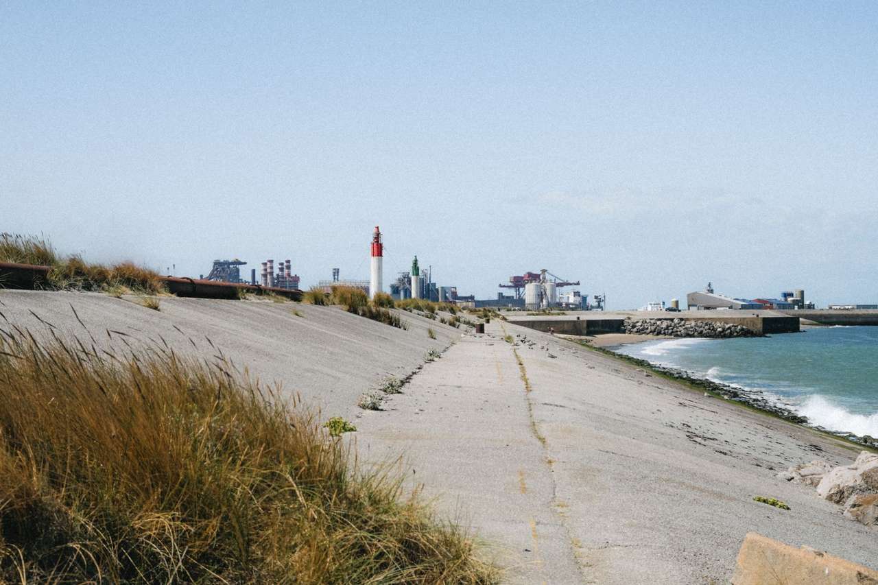 Digue en béton du littoral de Dunkerque, avec un feu de port et des installations industrielles et portuaires à l’horizon