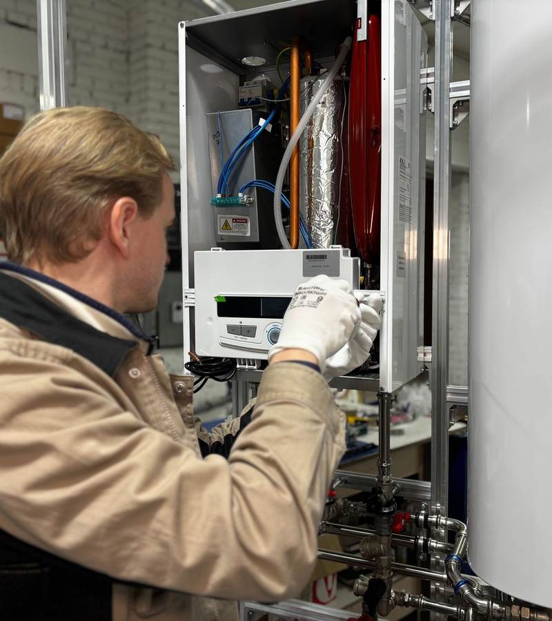 Technicien de dos, gants blancs, réglant une chaudière murale ouverte dans un atelier