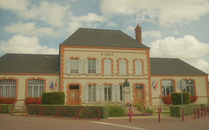 Façade d’une mairie de village en brique et pierre avec drapeaux et massifs fleuris