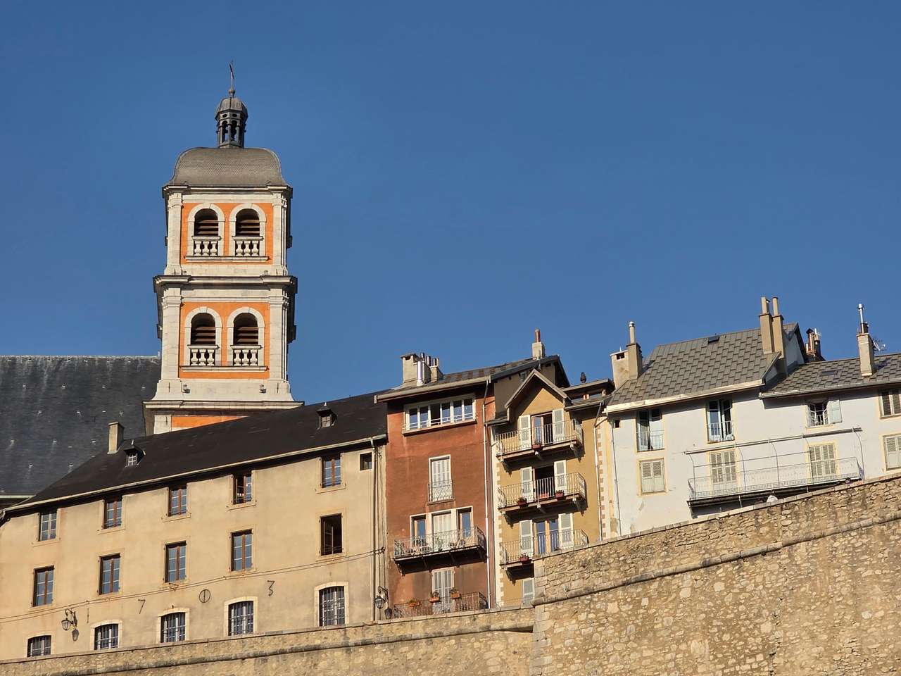 Clocher coiffé d’un dôme au-dessus d’immeubles anciens et d’un mur de pierre, à Chambéry