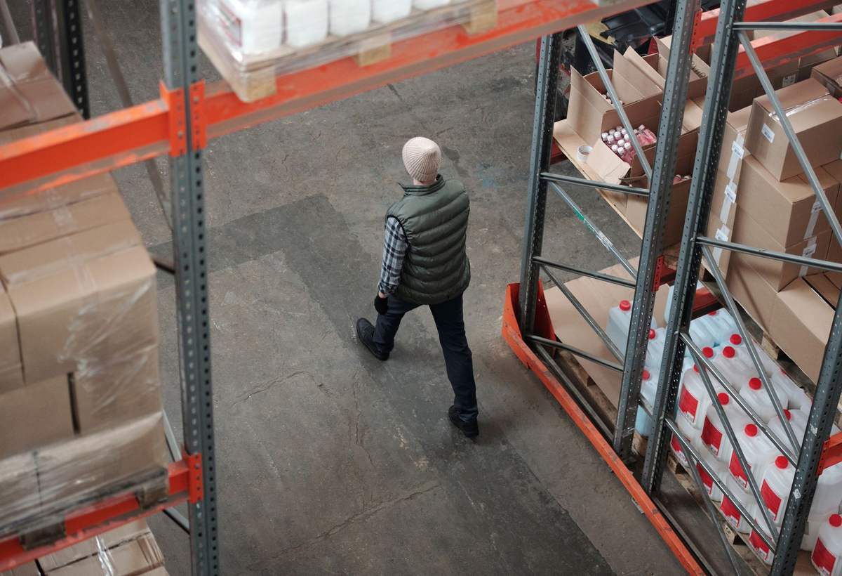 Homme en doudoune sans manches marchant entre des rayonnages de cartons dans un entrepôt, vu de haut