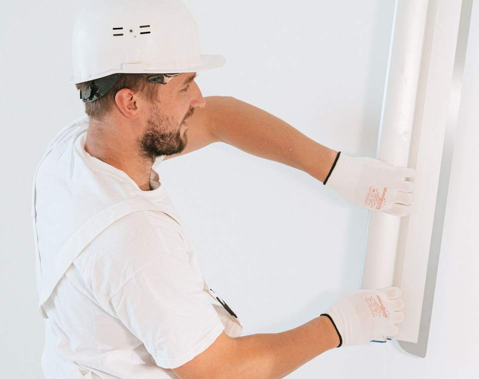 Ouvrier en casque blanc et gants de protection posant un profilé contre un mur blanc
