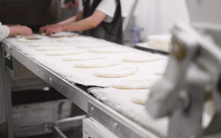 Pâtons aplatis sur un tapis fariné d’une ligne de boulangerie industrielle, opérateurs en arrière-plan