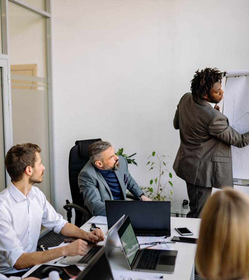 Formateur écrivant sur un paperboard devant des adultes assis autour d’une table avec ordinateurs portables