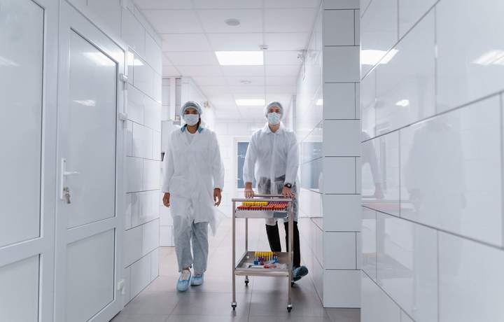 Personnel en blouse, charlotte et masque poussant un chariot de tubes de prélèvement dans un couloir carrelé